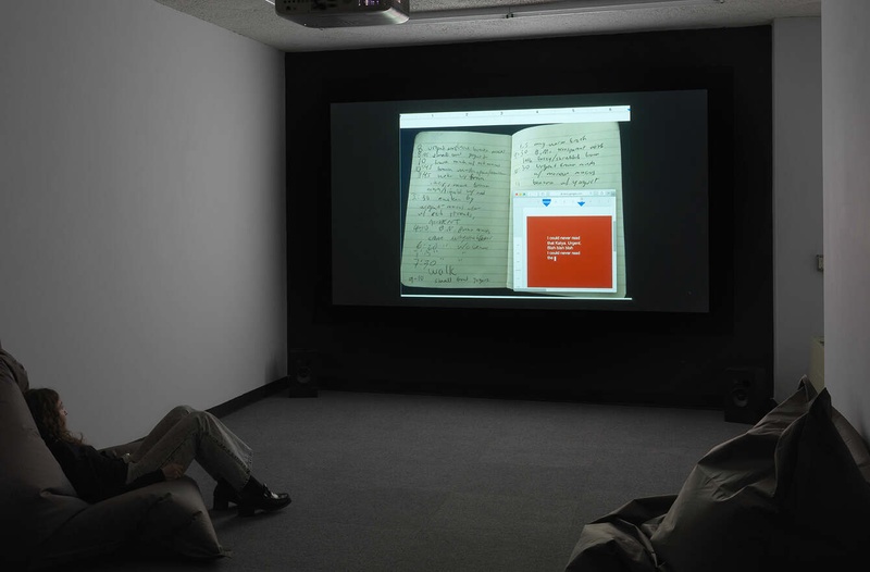  A projector in a dark room with two white walls and a grey floor with two beanbags, one occupied by a person with long hair and heeled boots. On the blackwall opposite the camera is a projection of a picture of two notebook pages. 