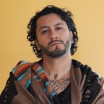 A headshot of a Latinx person of Mexican-American descent wearing a serape poncho posing in front of a yellow wall. His face is slightly tilted back, he is wearing a gold septum piercing and he is staring directly at the viewer.