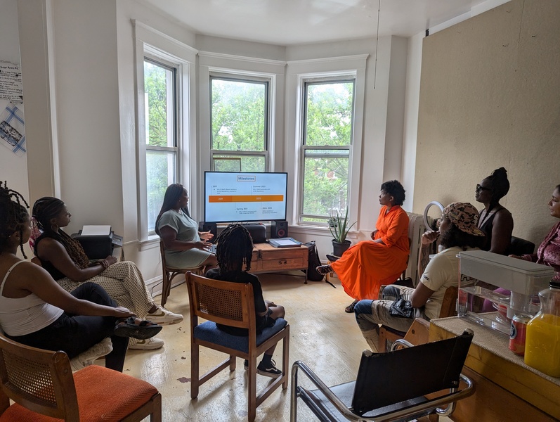  An audience listens as two people speak in a room with big bay windows. They are both sitting, one wears a green dress the other wears an orange dress. 