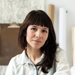 Mexican-American woman with brown hair and brown eyes, wearing a blue jean button down shirt sitting in her studio