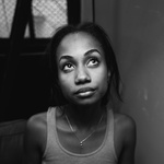 Black and white photo of artist looking up. Artist is a black woman with a medium complexion and straight black hair and brown eyes. She is wearing a gray tank top.