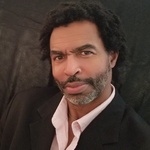 Black man with curly black-ish hair, brown eyes, and a Covid beard