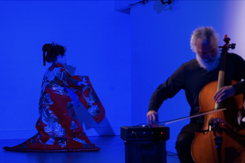  An Asian American woman dressed in traditional Japanese kimono, cast in a blue light, next to a person playing a cello. 