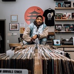 A headshot of a black/queer person in black and white. They have a wrap on their head and over ear headphones dangling from their head.