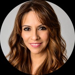 A Latino woman with long dark brown hair and brown eyes in front of a turquoise wall; she uses she/her pronouns.