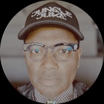 Sam is a Black man with glasses, wearing a black hat, a checkered shirt with a turquoise bolo tie, and a black vest. He's sitting in an empty theater. Photo by the artist.