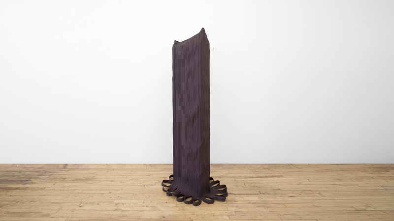  A sculpture of multiple zipper lines covering a wooden rectangle. The zippers land on the floor and curl. 