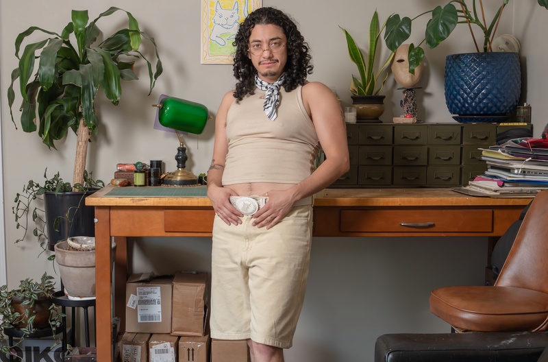  A person poses while leaning against a wooden desk. They have long brown curly hair and are holding on to their belt buckle and wearing an all tan outfit. 