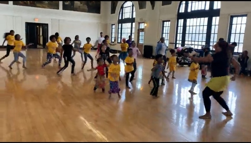  A group of 20 children dancing in a gymnasium, following the directions of instructor T. Ayo Alston at the front of class 