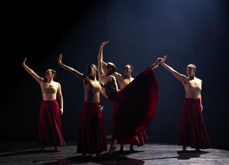  A group of five dancers performer in an all black room. They each wear long red skirts and tan bras. One dancer is posed with their leg in the air while the others right the hands up. 