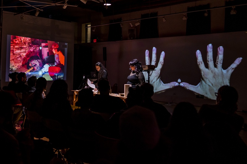  Two performers wearing black are standing in a dark room there is a projection of two hands behind them. 