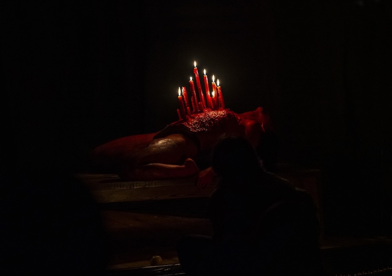  A performer lies on a table in the dark. She has long red lit candles melting on her abdomen. 