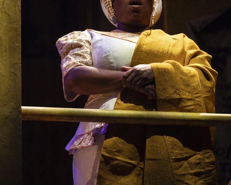  A close up of a costume on stage. It is a white pant and matching top with stylized patterned sleeves and collar. The performer also has a yellow wrap that they are holding on to. 