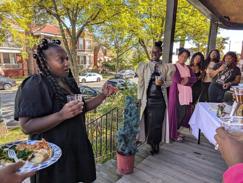  An artist wearing black with black braids talks to an audience outside on a porch during an artist reception. 