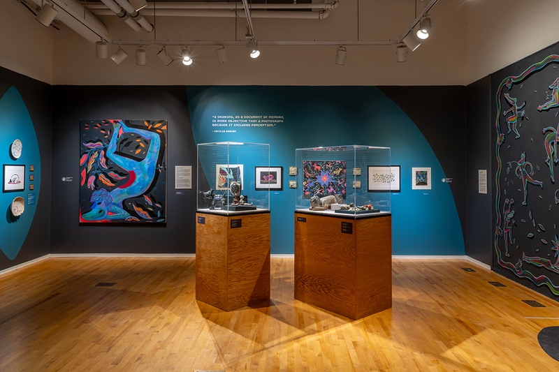 installation showing colorful paintings, drawings, ceramics, and a mural at a gallery with dark walls. The artwork shows animal-human hybrid figures 