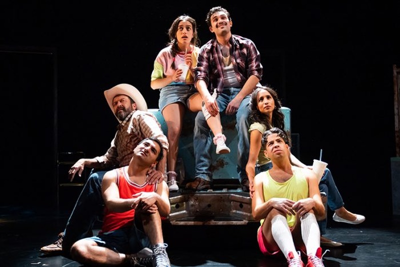  A still image from the play Rust. Six actors sit in a pyramid on stage while looking up towards the sky. They are all wearing different athletic wear and some western themed clothing. Jocelyn holds a straw and cup. 