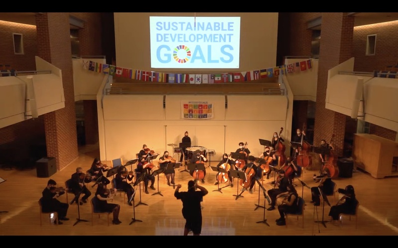  Ayriole, a brown skinned Black person with their back to the camera, on a low lit stage conducting a string ensemble. A string of international flags hang above the stage and a projected image reads "sustainable development goals" 