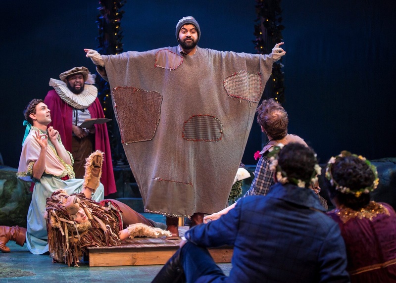  Richard wearing a patchwork blanket with arms outstretched, he has on a gray knit cap and a full beard. 
