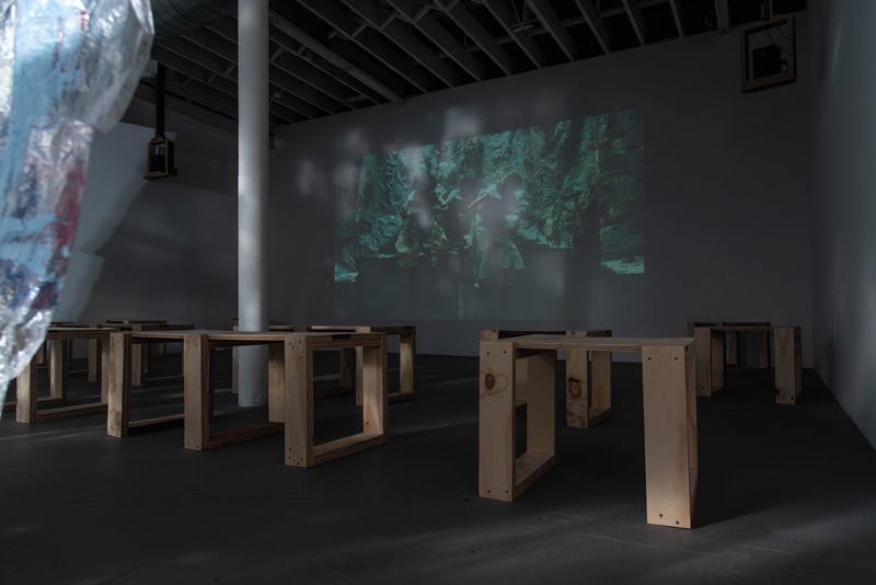  A dark room with brown wooden tables sit in front of wall with projected image of people 