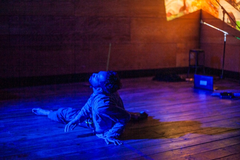  Light-skinned Black person with short black curly hair poses and they dance on wooden floor in a blue and yellow lit room. 