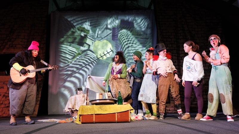  A group of performers dressed as clowns stand on stage behind them is a projection of distorted buildings and telephone polls. 
