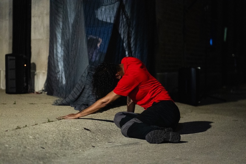  Light-skinned Black person with short black curly hair poses and they dance on a concrete floor outside a gallery. They are wearing a red shirt. 