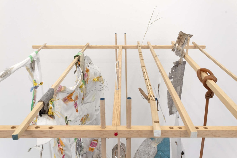  A close up of an art piece composed of pieces of lightly colored wood put together to make an angular structure. Seven pieces of wood lay between two parallel horizontal pieces, held up by two sets of two wooden rods, one on each side. 