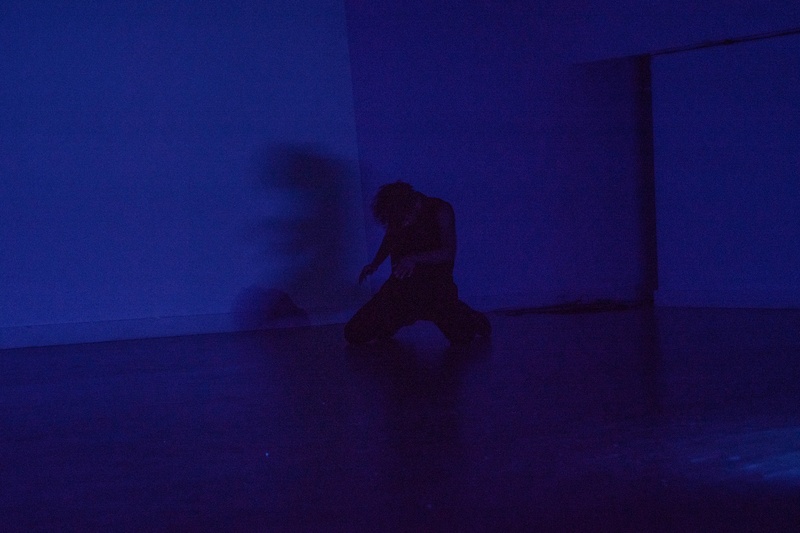  Light-skinned Black person with short black curly hair poses and they dance in an dark blue room. 