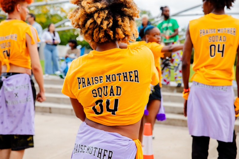  Kopano (PRAISE MOTHER SQUAD dancer) faces away from the camera showing off their yellow shirt with print of "PRAISE MOTHER SQUAD 04" 