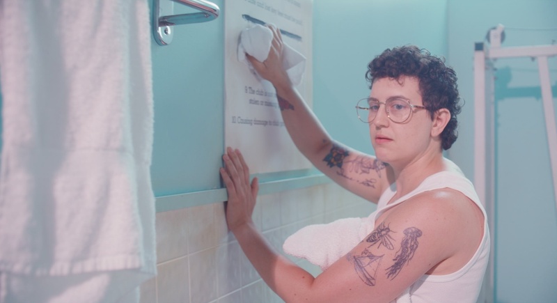  White, Queer, Non-binary Person with curly hair Glasses cleaning a sign on a blue wall in a bathroom. 