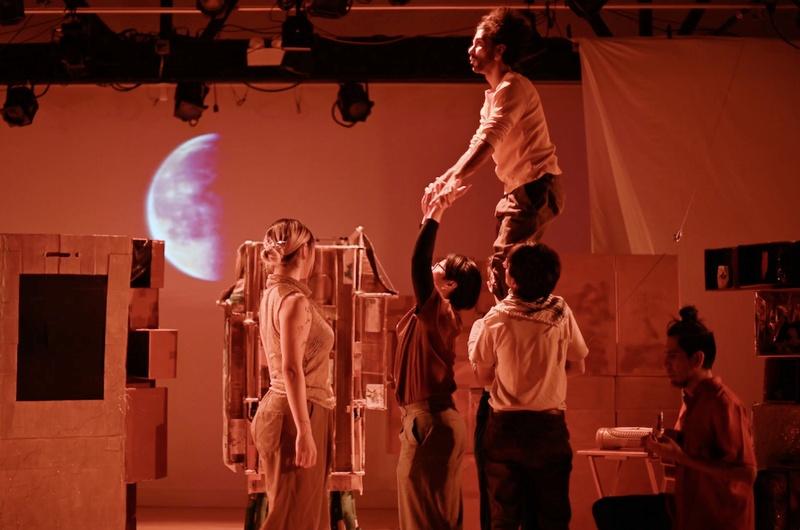  Three performers are lifting a four performer into the air in an orange lit room. Behind them is a projection of the moon. 