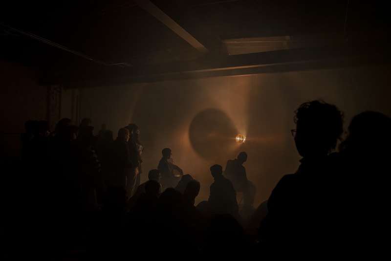  An audience watches shadows of performers in front of a low lit wall. 