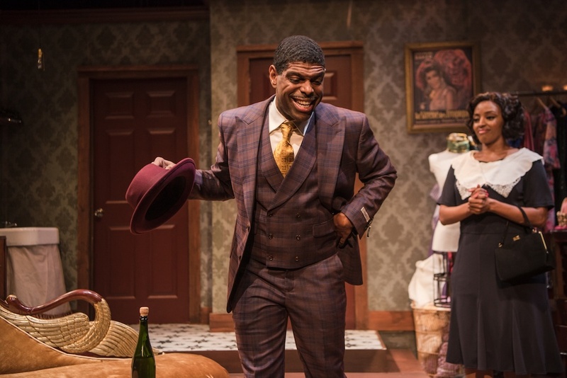  A still from a theater production. A Black, Queer performer stands in the frame in a pink and purple gingham suit. They are smiling and holding a pink hat. 