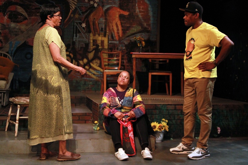  Three performers post on stage. On sits on the ground while the other two stand over them talking. 