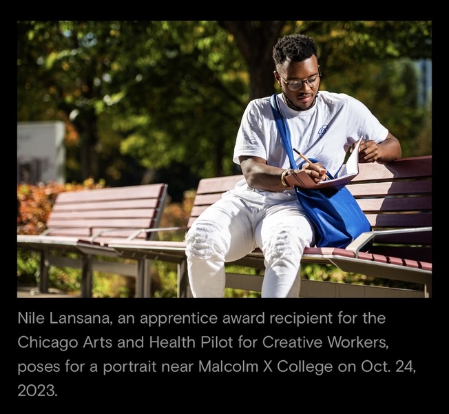  A Black Masc Person with short black locs and brown eyes sits on a park bench. He is wearing a light blue shirt and white pants. He is writing in a journal which is resting on a blue bag. 