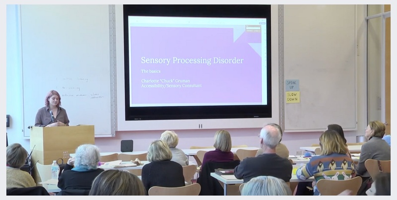  A person giving a presentation in front of a crowd of people. A presentation slide reads "Sensory Processing Disorder". 