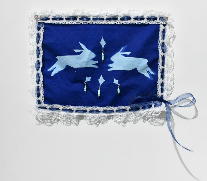  A blue textile hanging on the wall, in the middle are two rabbits facing each other with four diamonds stitched with beads. 