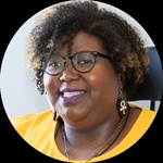 A close-up shot captures a smiling plus-sized Black woman with short, curly black hair that has blonde highlights. She is seated in a black wheelchair with a high headrest against a plain, light grey background.