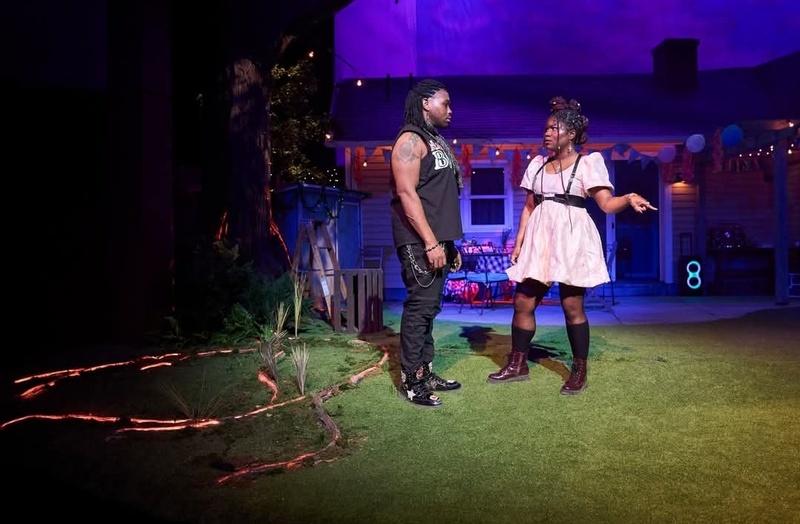  A production still from Fat Ham. Two performers are standing in a backyard with the open view of a living room. One performer is wearing a pink dress and black belt with suspenders. The other is wearing a black tank top, pants and cowboy boots. 