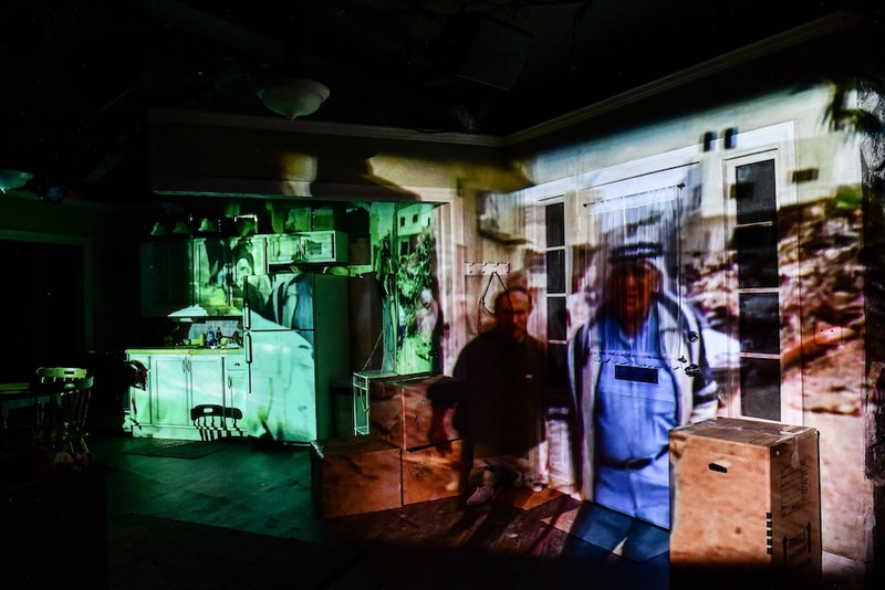  Projections of two man standing outside displayed over the theater set of a kitchen in a home. 