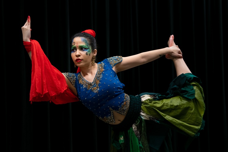  woman wearing colorful outfit dances while holding leg and pointing to the sky with her fingertips painted in red 