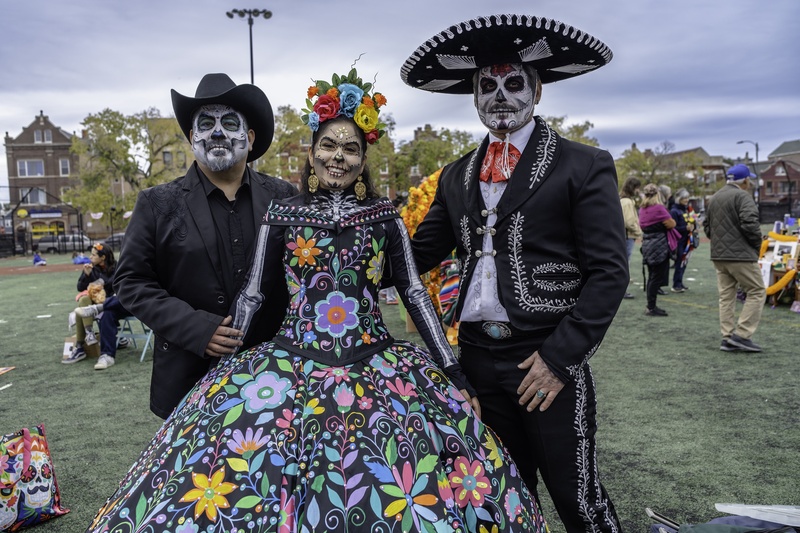  Three people pose for a photograph. They are wearing skeleton makeup on their faces and costumes styled for dia de los muertos. 