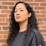 Korean American woman with dark curly hair falling over her shoulders and wearing a black top stands looking at the camera in front of a brick wall