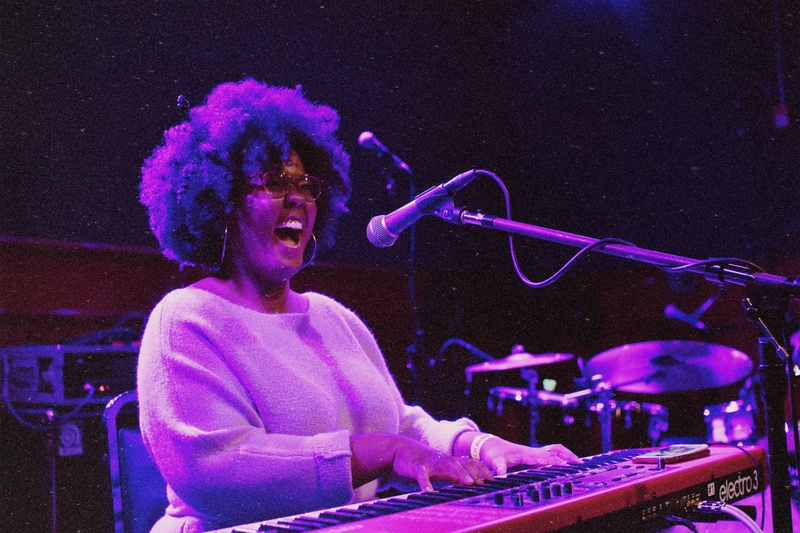  Black woman with curly hair and glasses, sits at a keyboard with mouth open in song. 