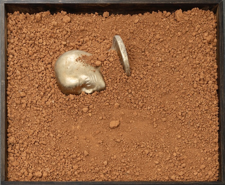  Image of a sculpture made with ray clay. There is a bronze bust in the clay dirt. Everything is contained in a black box. 