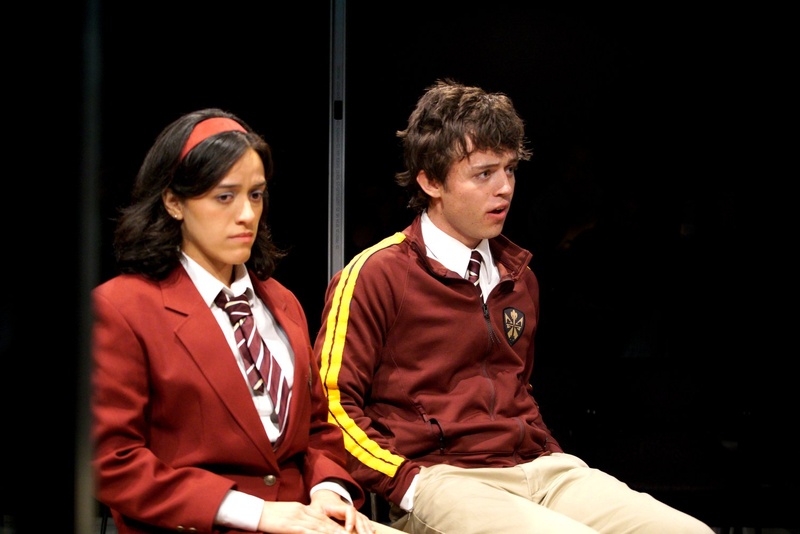  A still from the play Happy Days are Here (Again). Two people sit on a bench. One has a maroon school girl outfit on while the other has a maroon track jacket. 