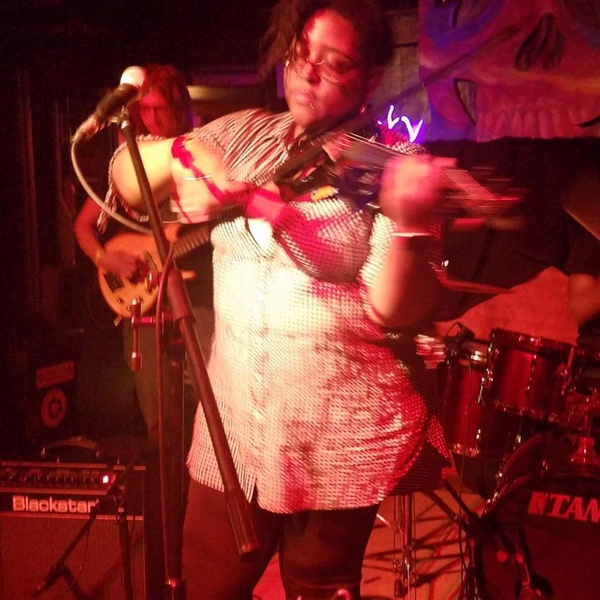  Photo of Ayriole, a brown skinned Black person, under reddish stage lighting, wearing a light-colored button-down shirt, playing their violin on a stage with other band members behind them 