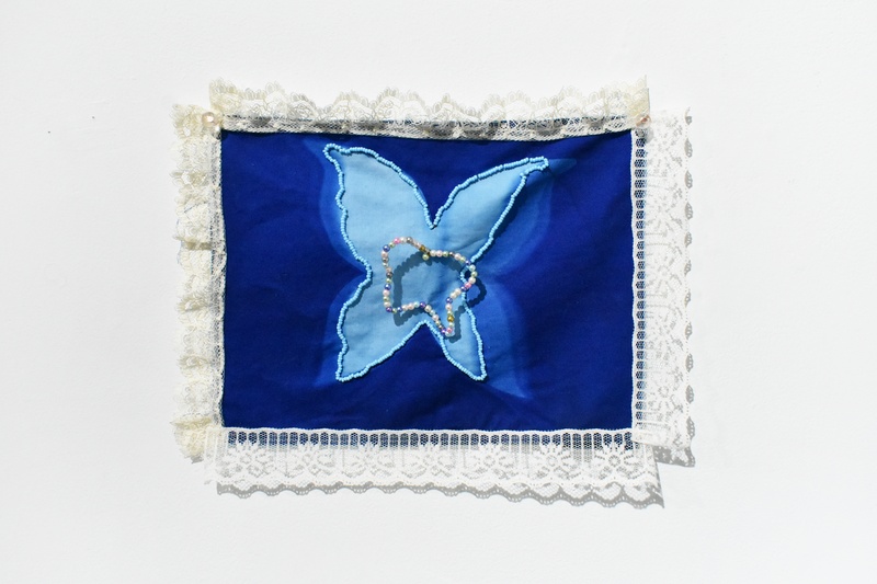  A cyanotype with varying shades of blue to mimic the wings of a butterfly fluttering, with a beaded butterfly in the center and a lace boarder 