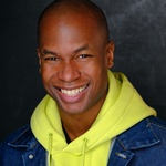 Black man smiles, wearing a yellow hoodie and denim jacket