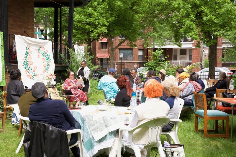  A group of people sit at white tables outside while observing someone read a story. 